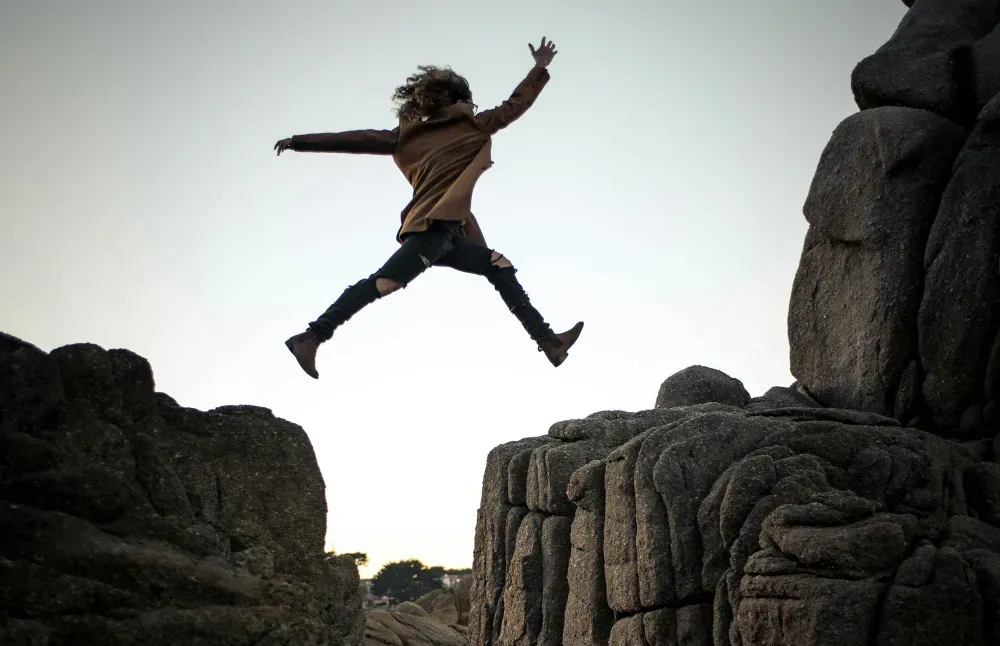 person jumping on big rock under gray and white sky during daytime